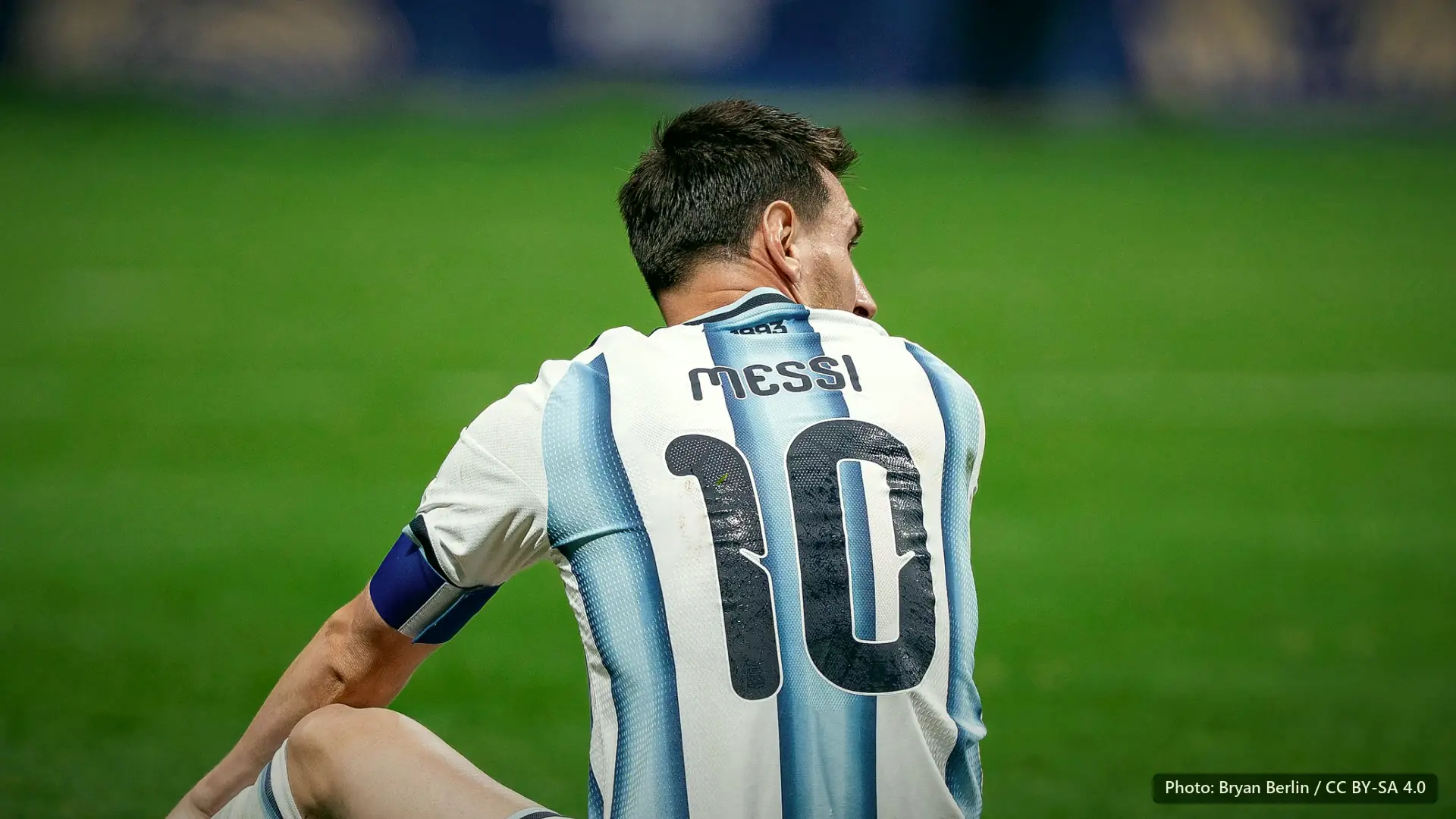 Messi seated on the grass, seen from behind and turning his head, in the Argentina number 10 shirt.
